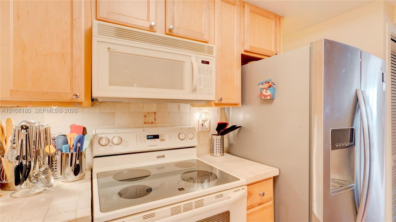 Light wood-finish cabinetry, white tile backsplash, integrated over-the-range microwave, smooth-top electric range, and stainless steel refrigerator