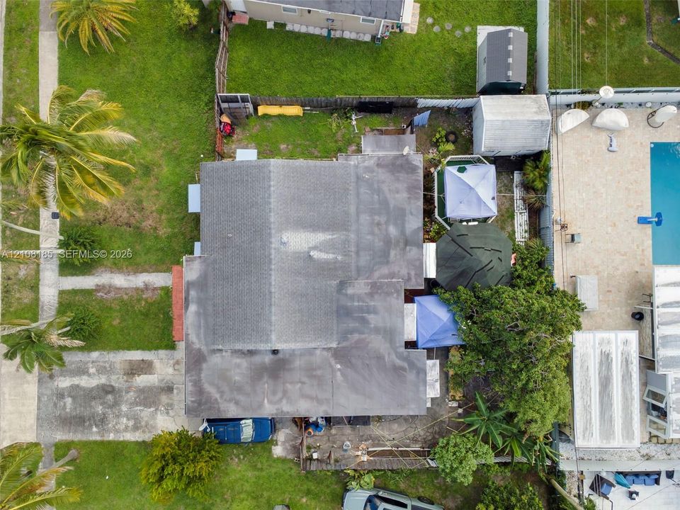 Aerial perspective of a residential lot featuring a multi-section asphalt shingle roof, paved driveway, expansive lawn, and established palm trees
