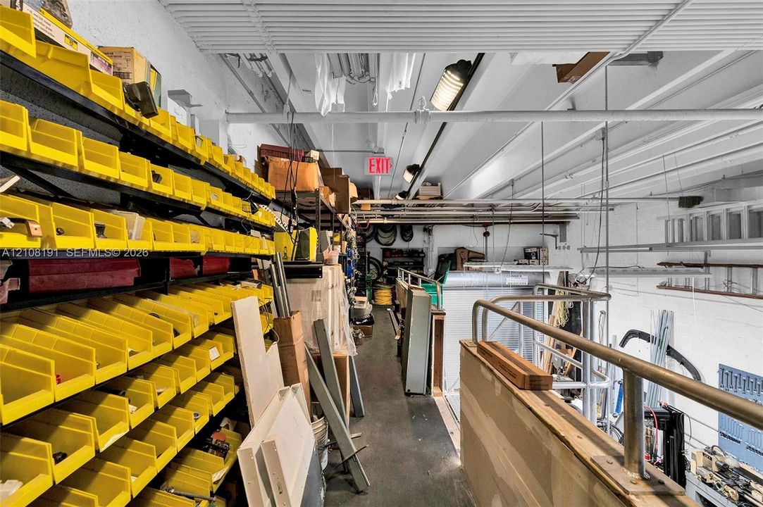 Upper-level storage mezzanine featuring integrated wall-mounted bin shelving, exposed industrial ceiling, and a metal safety railing overlooking the lower floor