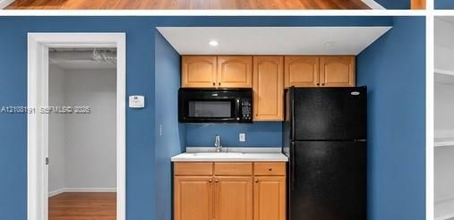 Kitchenette featuring light wood cabinetry, integrated microwave, white countertop with sink, and black refrigerator