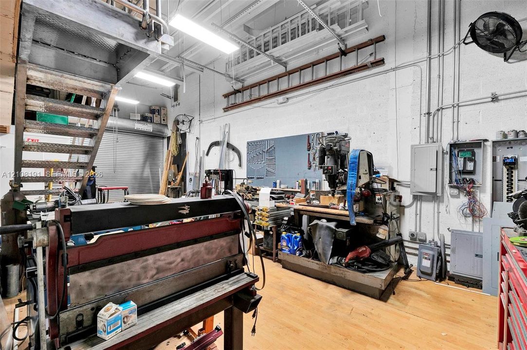 Industrial workshop space featuring high ceilings, exposed structural elements, steel stairwell, concrete walls, and roll-up metal door