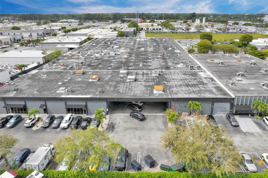 Industrial commercial property featuring a flat roof with roof-mounted HVAC units, multiple ground-level overhead doors, expansive paved surface parking, and exterior metal siding