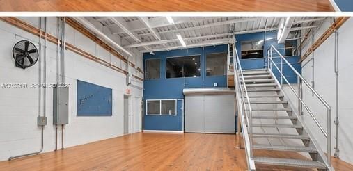 Open-concept interior featuring wood-finish flooring, exposed ceiling beams, a metal industrial staircase, roll-up door, and elevated interior windows