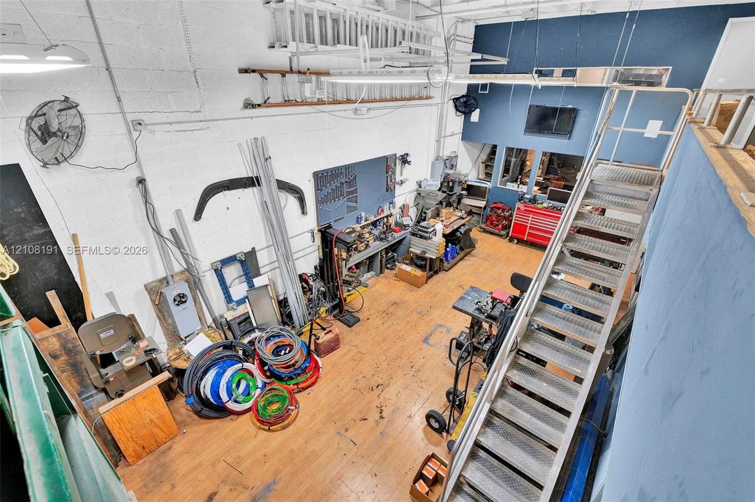 High-ceiling workshop featuring wood-finish flooring, industrial metal staircase, exposed structural elements, and built-in task lighting
