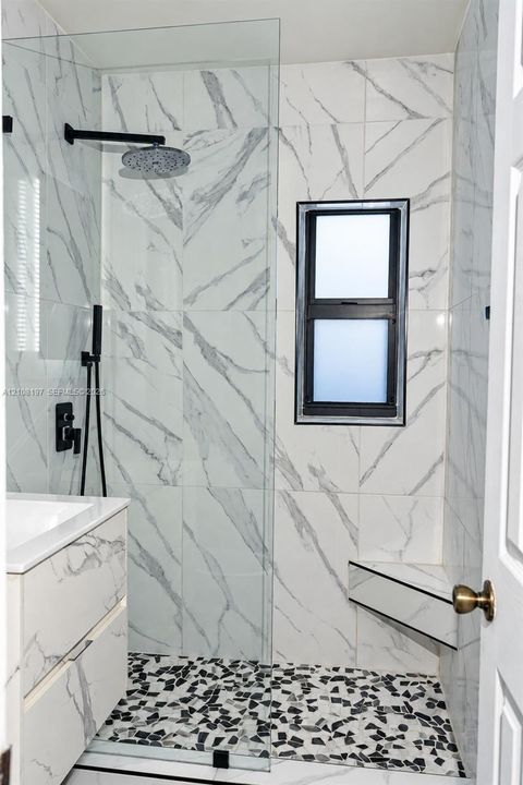 Walk-in shower featuring large-format stone-patterned wall tile, a built-in corner seat, matte black rainfall showerhead with handheld attachment, pebble-texture flooring, and a frameless glass partition