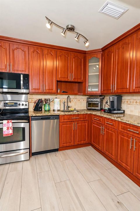 Kitchen featuring raised-panel cherry wood cabinetry, speckled stone countertops, stainless steel appliances, decorative tile backsplash, and wood-finish flooring
