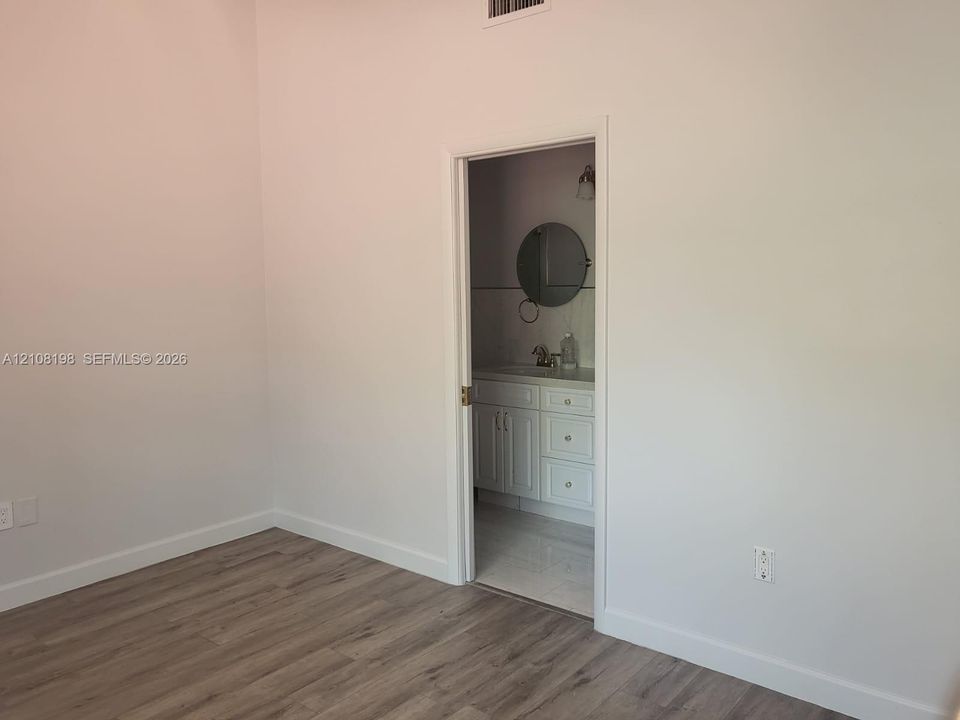 Room featuring wood-finish flooring and a doorway leading to a bathroom equipped with a white vanity cabinet, light-colored countertop, and a circular wall-mounted mirror