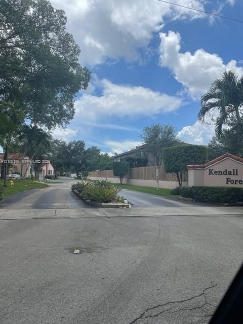 Community entrance featuring masonry signage, mature trees, manicured hedges, and palm landscaping