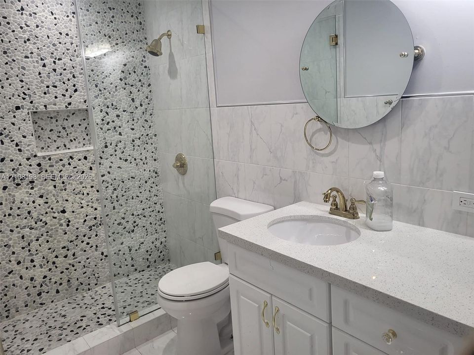 Bathroom featuring a glass-enclosed shower with a pebble-tile accent wall, built-in shower niche, stone-composite vanity top with an integrated sink, and a circular wall-mounted mirror