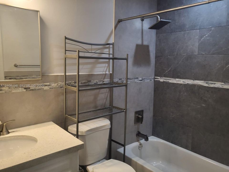 Bathroom featuring a vanity with a light-toned solid surface countertop, rectangular mirror, glass and stone mosaic tile backsplash, and a tub-shower combination with large-format dark tile surround and a rainfall showerhead
