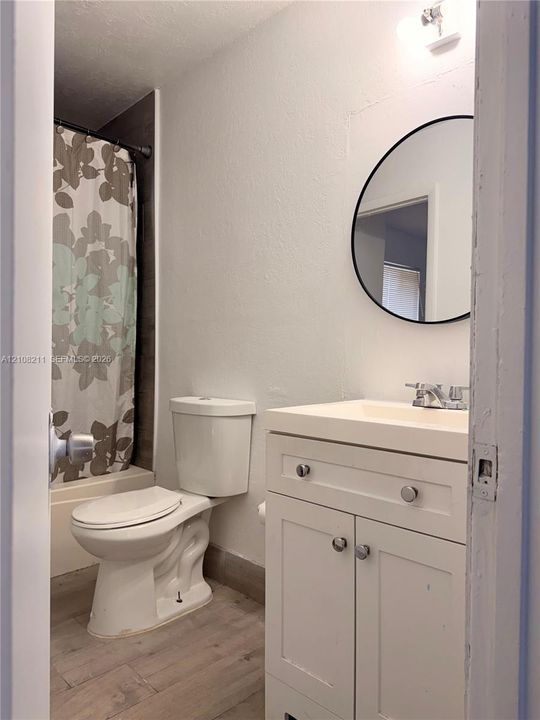 Bathroom featuring a vanity with white cabinetry, a circular wall-mounted mirror, wood-finish flooring, and a shower-tub combination