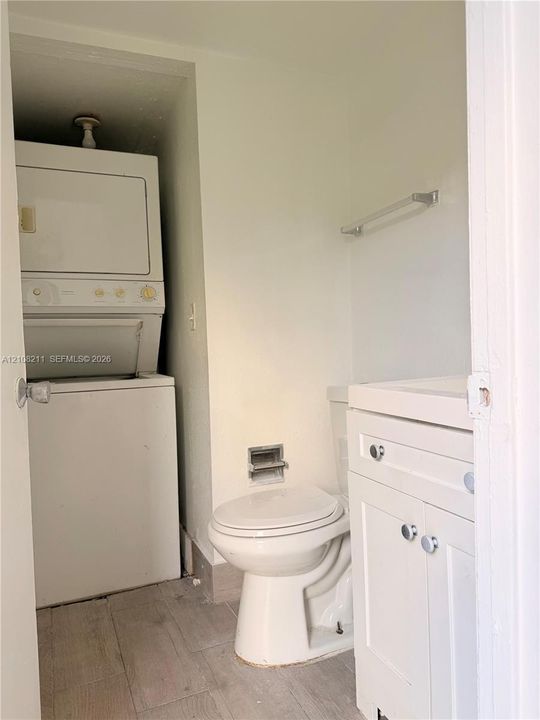 Stacked laundry unit, white vanity cabinet with a rectangular basin, ceramic toilet, and wood-finish flooring