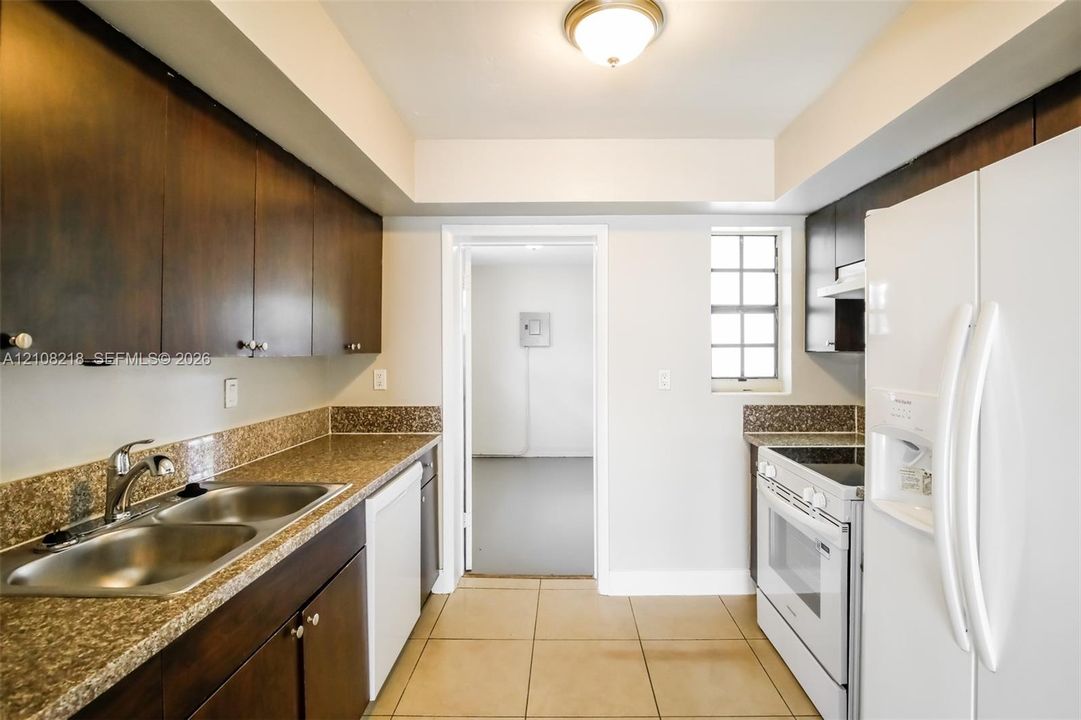 Kitchen featuring dark wood-finish cabinetry, stone-surface countertops, double-basin stainless steel sink, ceramic tile flooring, and integrated white appliances