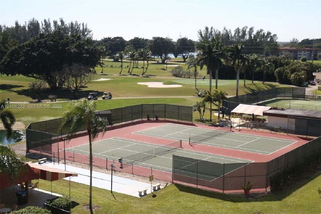 Dual fenced tennis courts featuring hard-surface playing areas, accompanied by a covered pavilion, set against a backdrop of a golf course with mature landscaping and water features