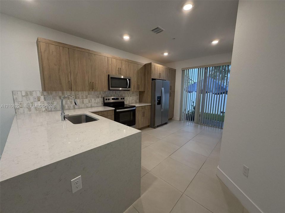 Kitchen featuring wood-finish cabinetry, quartz countertops, a stone-tile backsplash, stainless steel appliances, and large-format tile flooring
