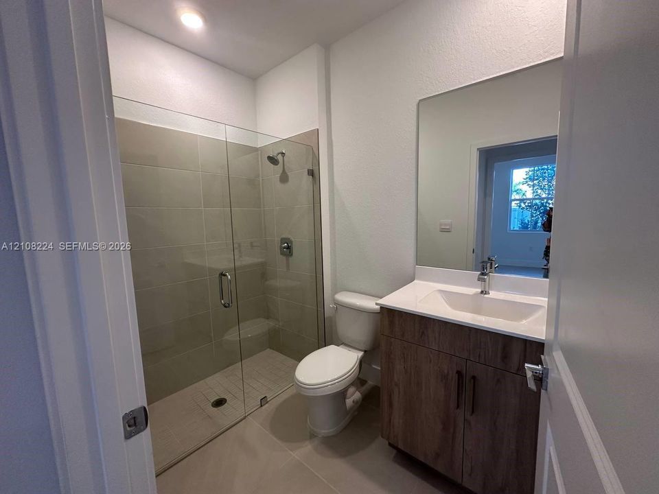 Walk-in shower with floor-to-ceiling tile, wood-finish vanity with an integrated basin, countertop with stone-style surface, and frameless vanity mirror