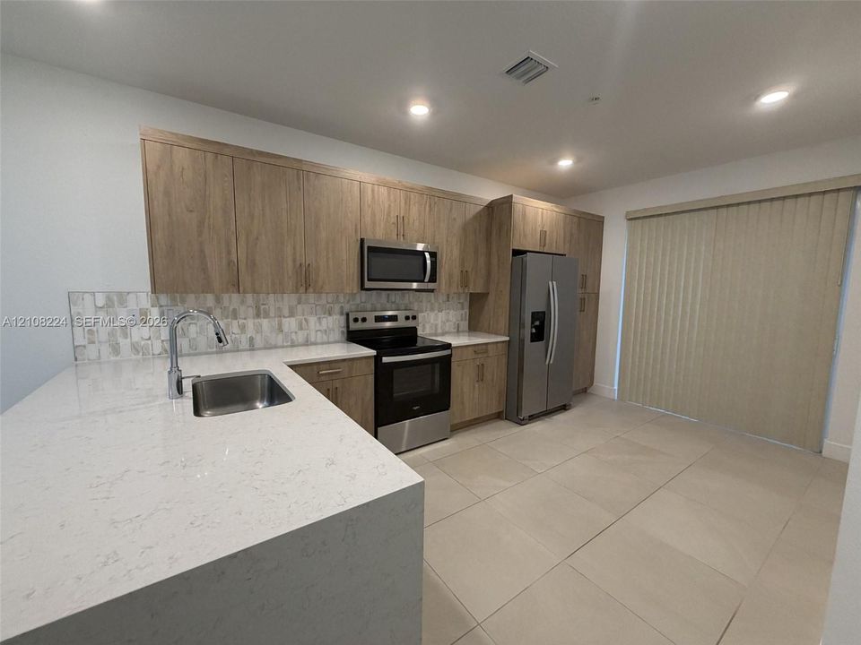 Kitchen featuring wood-finish cabinetry, quartz countertops, a stone-tile backsplash, stainless steel appliances, and large-format tile flooring