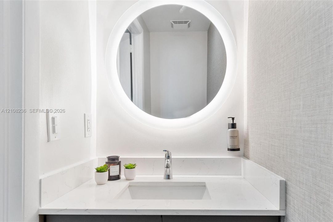 Circular backlit wall mirror, stone-composite countertop with integrated sink, chrome single-lever faucet, and textured wall finish
