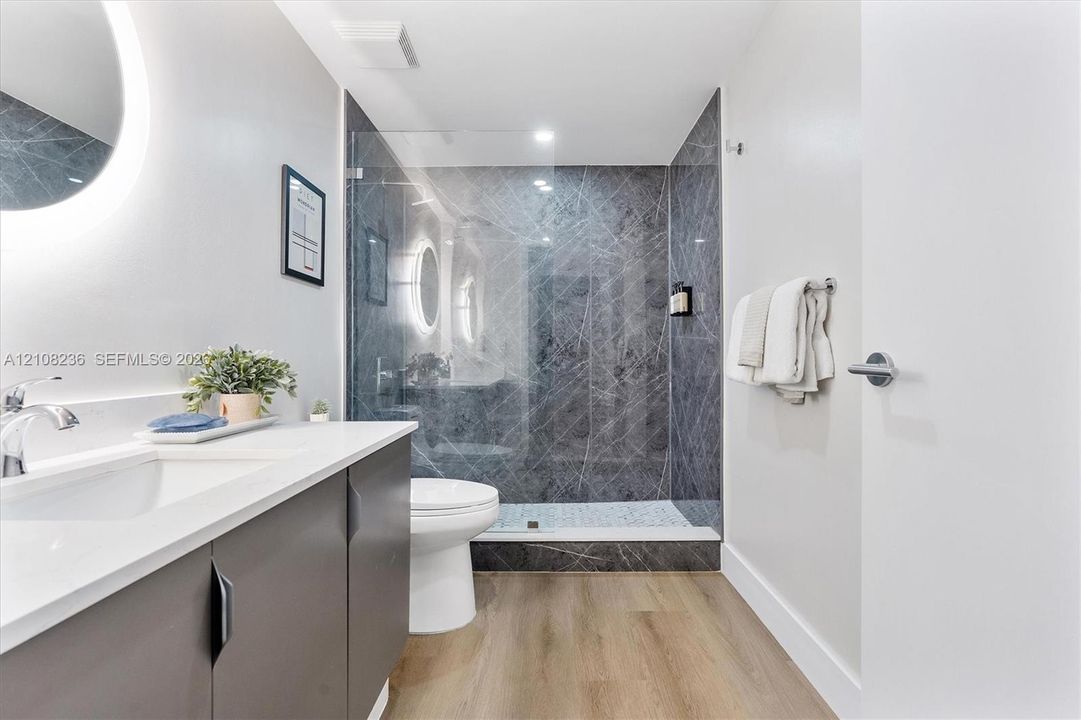 Modern bathroom featuring a dark vanity with a white countertop, walk-in shower with stone-patterned tile surround, glass enclosure, backlit circular mirror, and wood-finish flooring