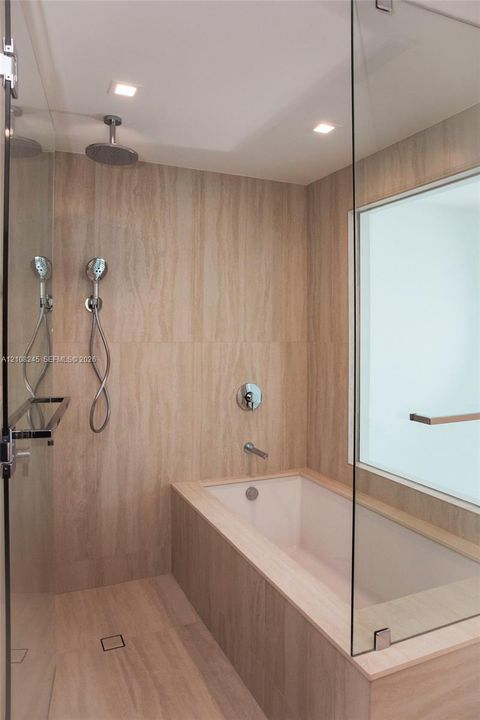 Walk-in shower and bathtub combination featuring floor-to-ceiling stone-tiled walls, glass enclosure, polished chrome rainfall shower head with handheld attachment, and recessed lighting