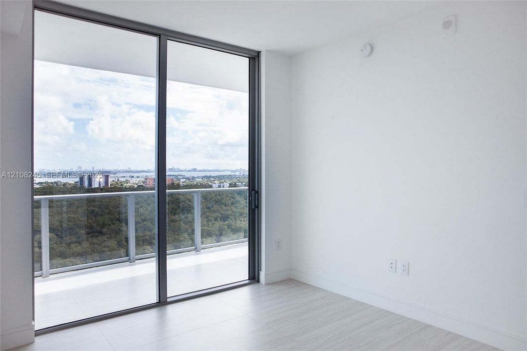 Floor-to-ceiling glass sliding doors opening to a private balcony with a glass railing and expansive horizon backdrop, featuring light-toned flooring and neutral wall finishes
