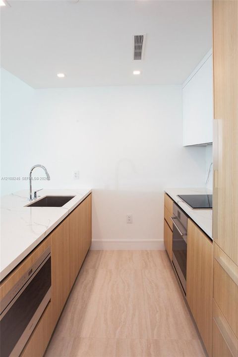 Kitchen galley featuring light wood-finish cabinetry, white stone-finish countertops, an undermount sink with a gooseneck faucet, a built-in oven, and a cooktop