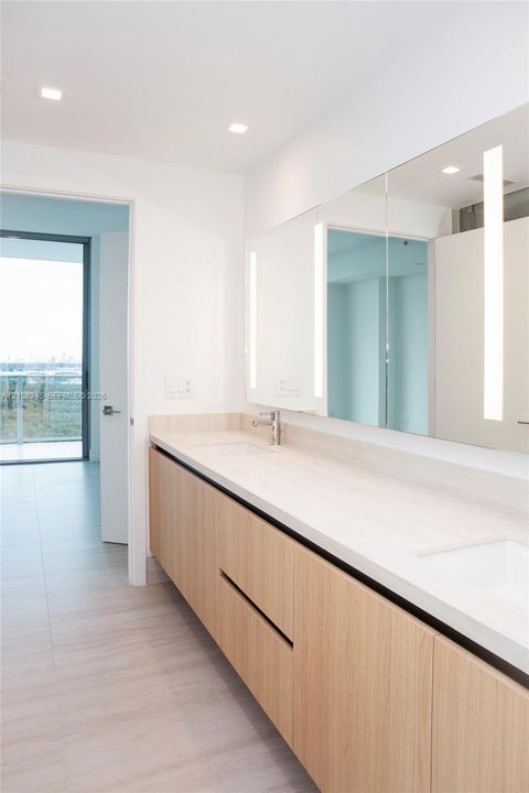 Floating vanity featuring a wood-finish cabinet, dual undermount sinks, stone-finish countertop, full-width mirror with integrated vertical lighting, and recessed ceiling lighting