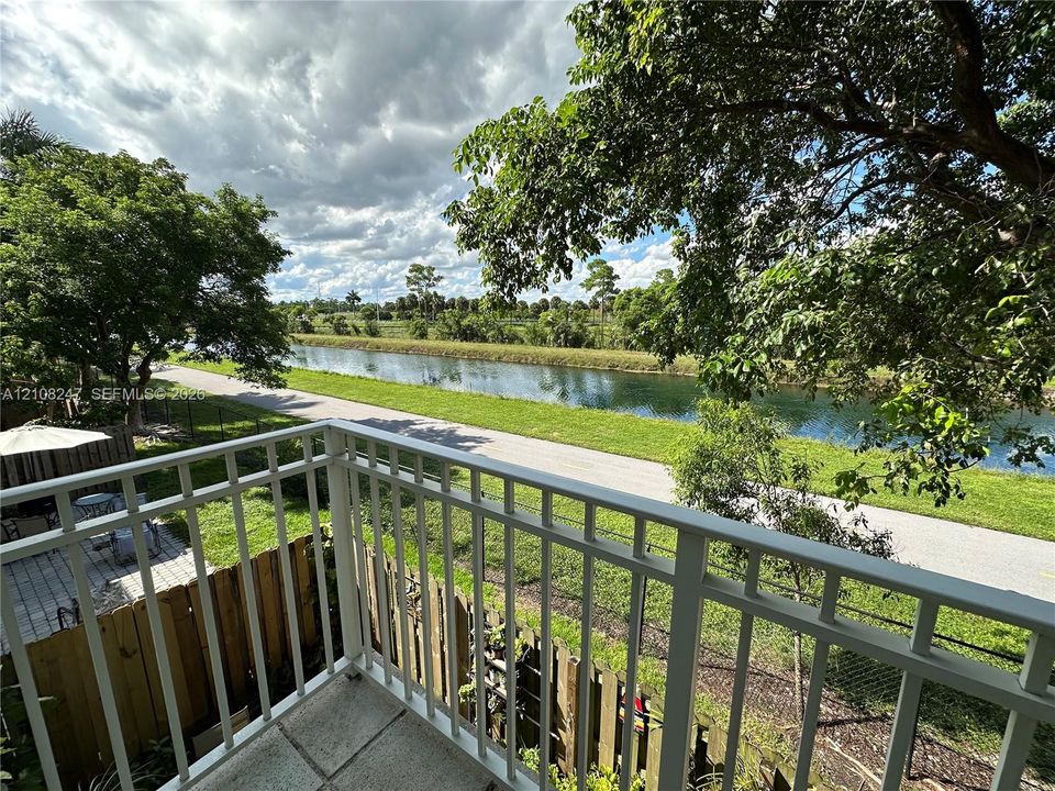 Elevated balcony with a white metal railing, paved patio area, wood fencing, and water frontage