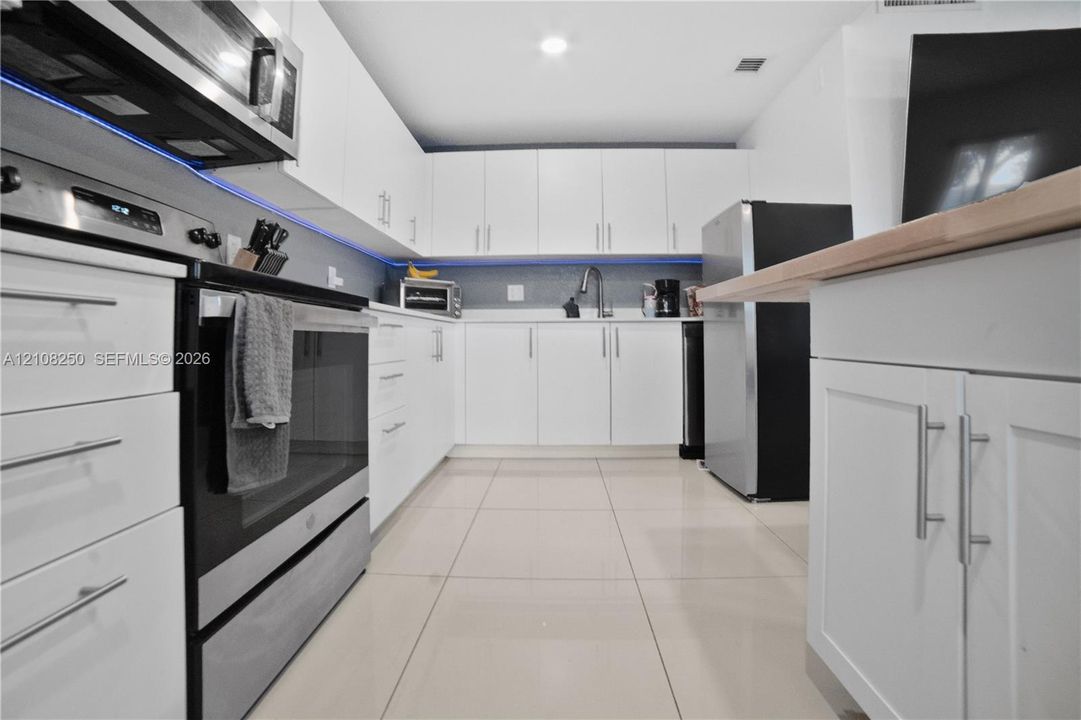 Kitchen featuring white flat-panel cabinetry, stainless steel appliances, tiled flooring, integrated under-cabinet lighting, and a wood-finish countertop