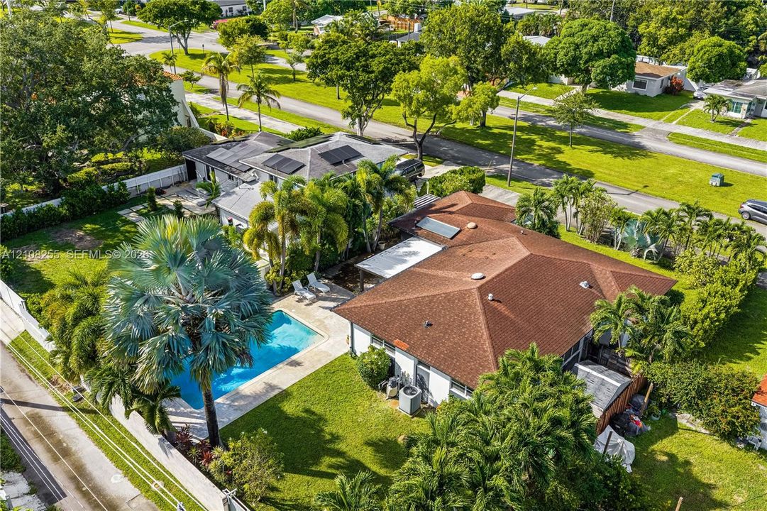 Aerial perspective of a property featuring an in-ground swimming pool, a patio area with a pergola, roof-mounted solar panels, and mature palm landscaping