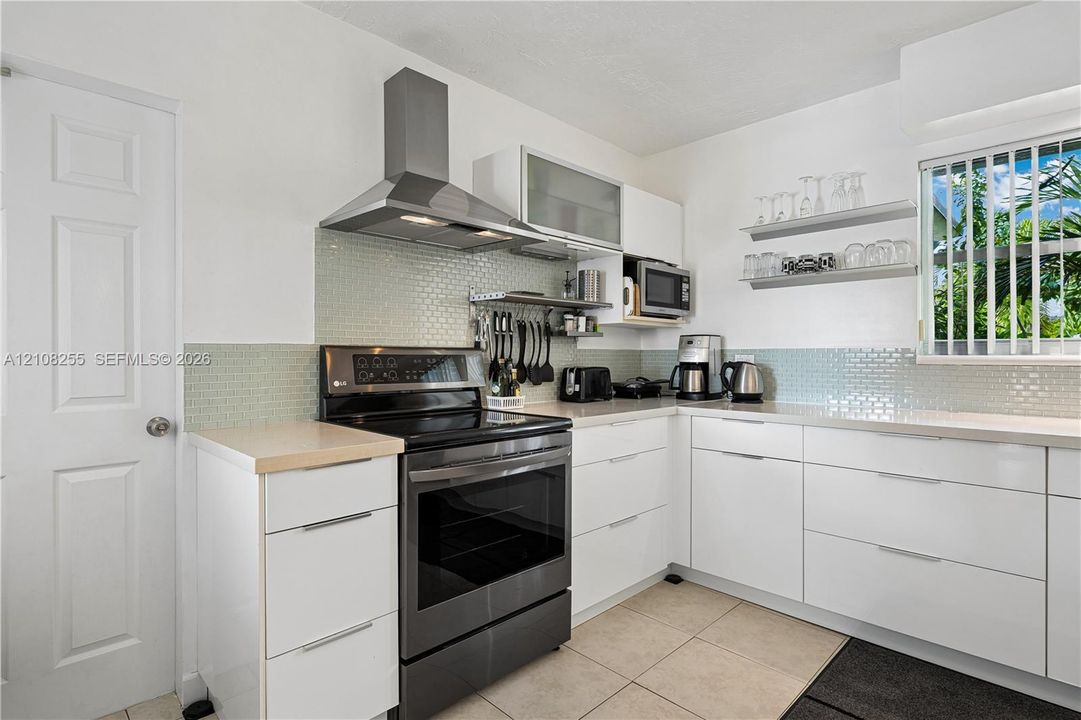 Kitchen featuring white flat-panel cabinetry, light-colored tile backsplash, stainless steel range with overhead ventilation hood, neutral countertops, and tile flooring
