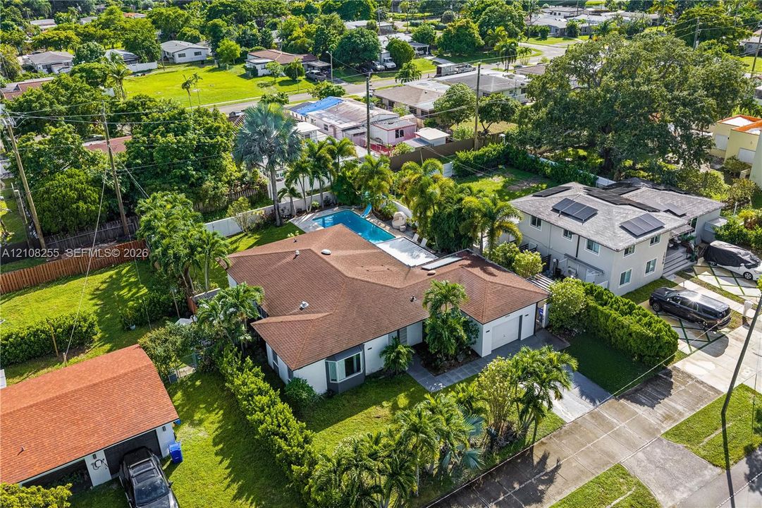 Aerial perspective of a single-story residence featuring a shingled hip roof, a private rectangular swimming pool, integrated solar panels, and manicured landscaping with mature palm trees