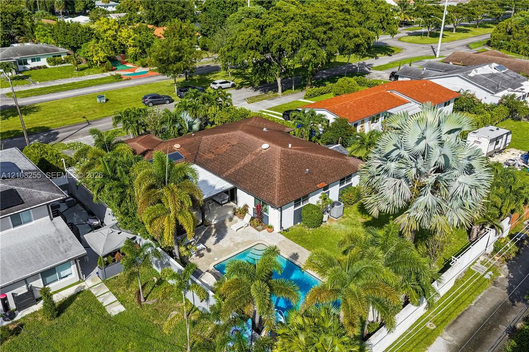 Aerial perspective featuring a shingle-roof residence, an in-ground swimming pool, a concrete patio, established palm trees, and a fenced perimeter