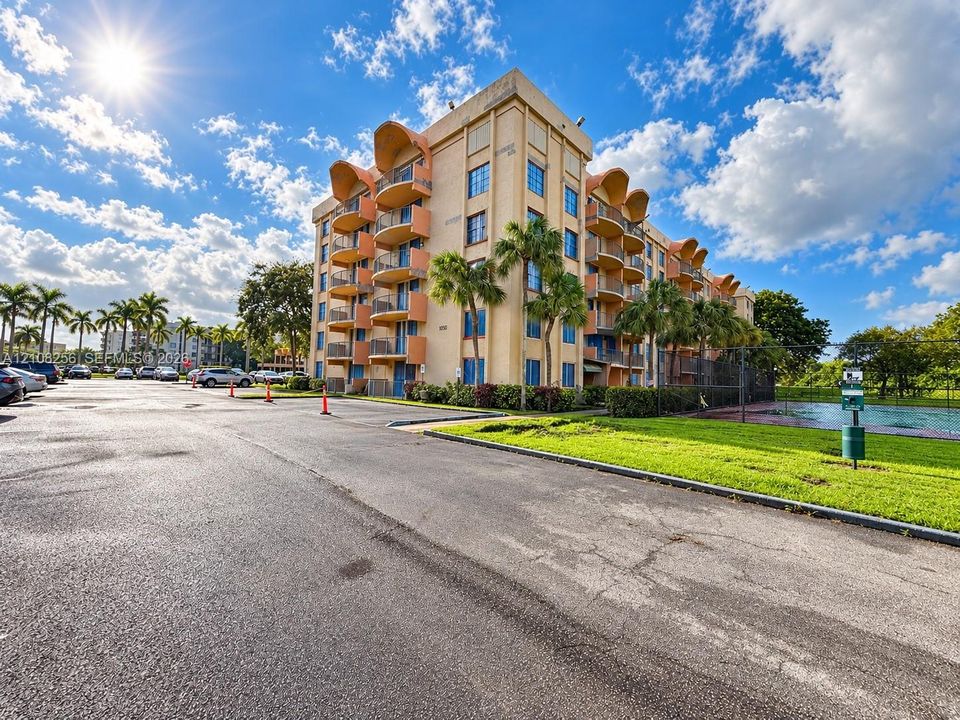 Multi-story stucco exterior featuring arched balconies, metallic railings, paved parking area, and landscaped grounds