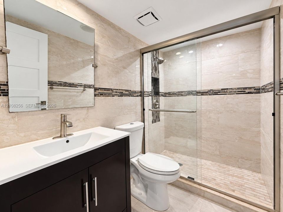 Bathroom featuring a dark wood-finish vanity with a white countertop, walk-in shower with sliding glass doors, neutral wall tiling with a mosaic accent border, and a frameless mirror