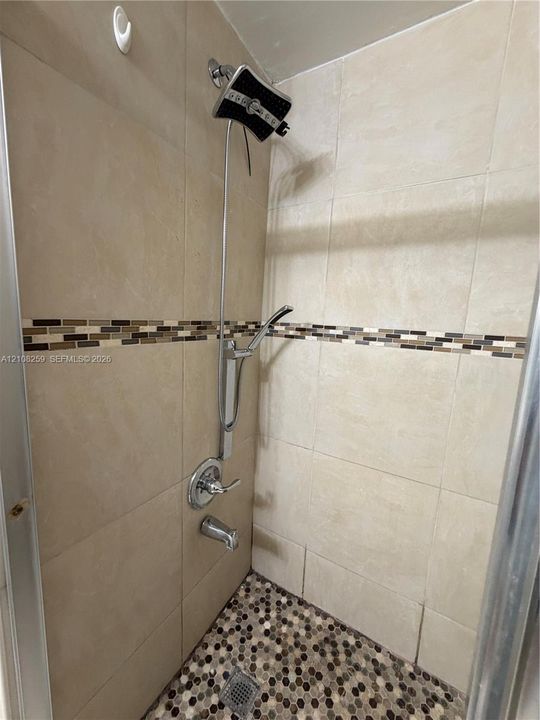 Shower featuring large-format beige wall tiles, decorative glass mosaic accent border, chrome rainfall shower head with handheld attachment, and multi-toned hexagonal tile flooring