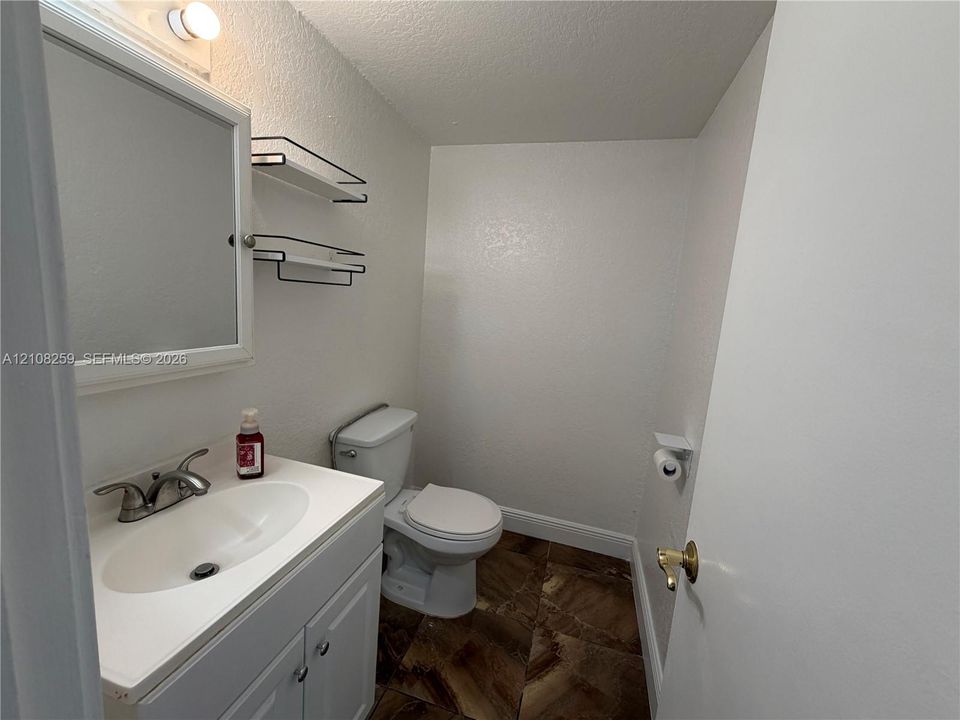 Bathroom featuring a white vanity cabinet with a neutral integrated countertop, double-handle faucet, wall-mounted medicine cabinet, dual metal shelving, and stone-finish tile flooring