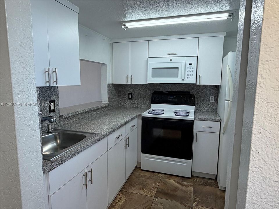 Kitchen featuring white shaker-style cabinetry, speckled stone-finish countertops, stainless steel sink with a high-arch faucet, integrated microwave, and tile flooring