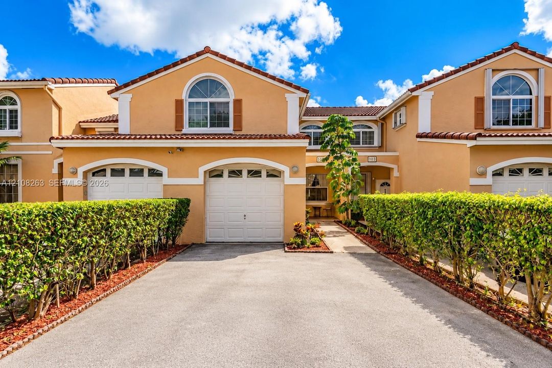 Stucco exterior featuring arched garage entryways, decorative shutters, clay tile roofing, and manicured privacy hedging