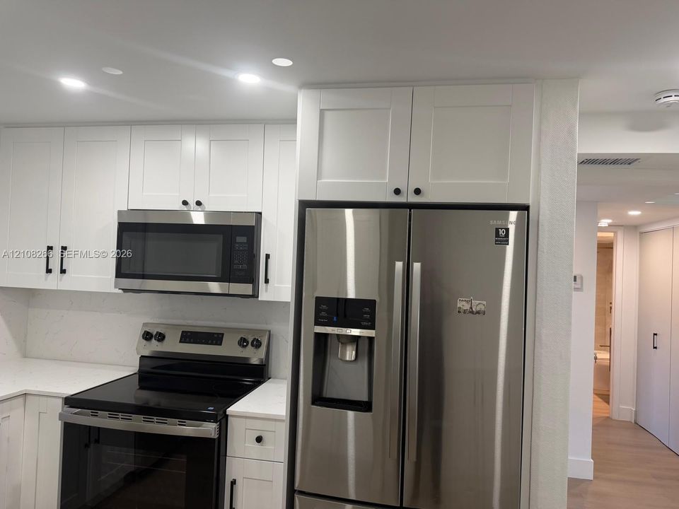 White shaker-style cabinetry with matte black hardware, stone-finish countertops, stainless steel side-by-side refrigerator, built-in stainless steel microwave, and recessed lighting