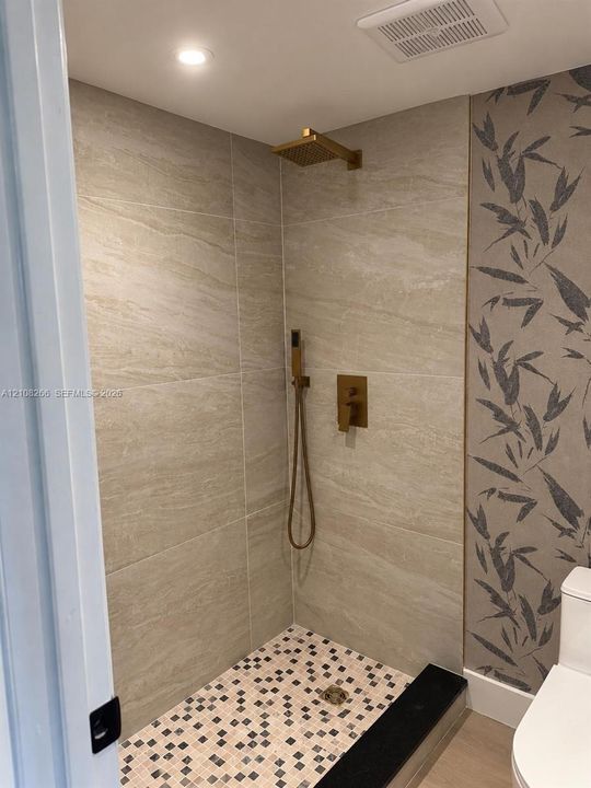 Walk-in shower featuring large-format stone-finish tile surround, mosaic floor tiling, gold-tone rainfall showerhead, and matching hand-held sprayer