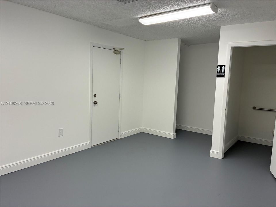 Interior space featuring gray seamless flooring, crisp white walls, a flush-mount rectangular light fixture, and an entry door with commercial-style hardware