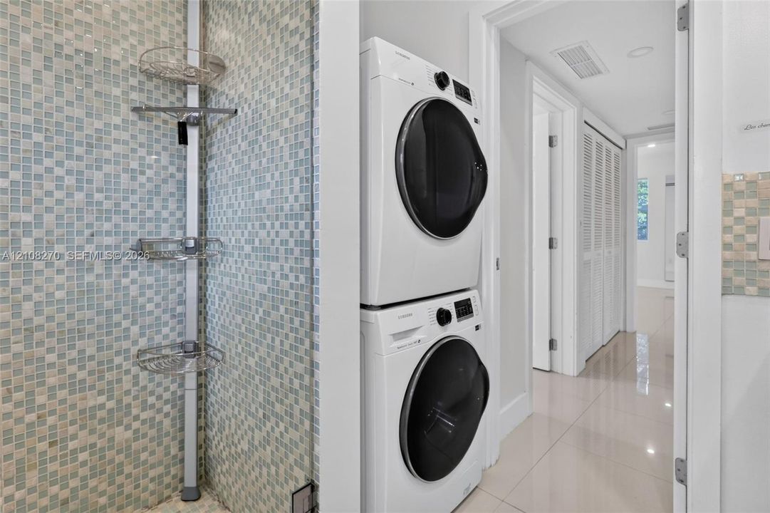 Laundry area featuring stacked front-load washer and dryer, tiled wall with integrated shelving, and polished neutral-tone flooring