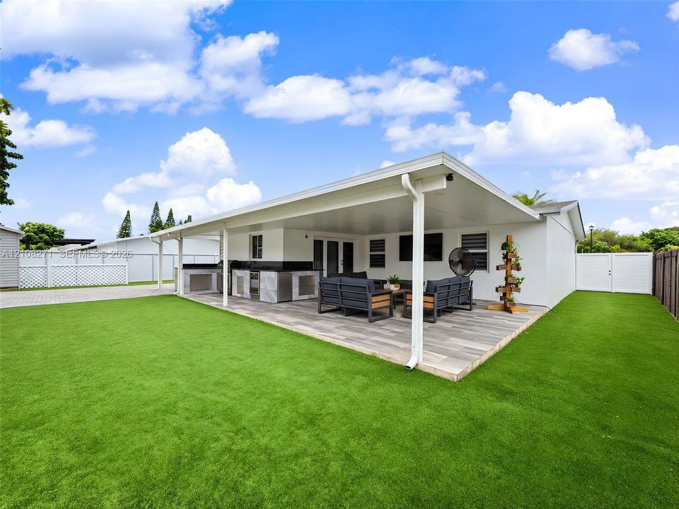 Covered patio featuring wood-finish tile flooring, integrated outdoor kitchen with stone veneer and dark countertop, and synthetic turf lawn