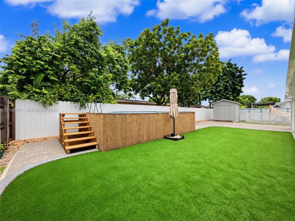 Backyard featuring an above-ground swimming pool with wooden stairs, synthetic turf lawn, paver patio, privacy fencing, and a detached storage shed