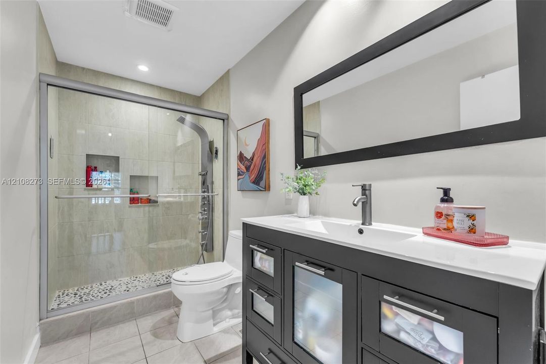 Dark-finish vanity with white countertop and rectangular integrated sink, walk-in shower with stone tile flooring and recessed shelving, and a large rectangular mirror with a black frame