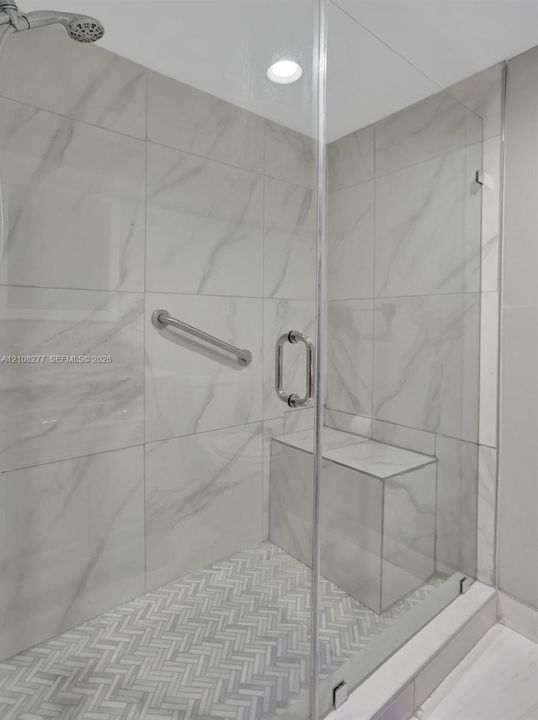 Walk-in shower featuring large-format porcelain wall tile, herringbone-patterned floor tile, frameless glass enclosure, built-in tile bench, and polished chrome hardware