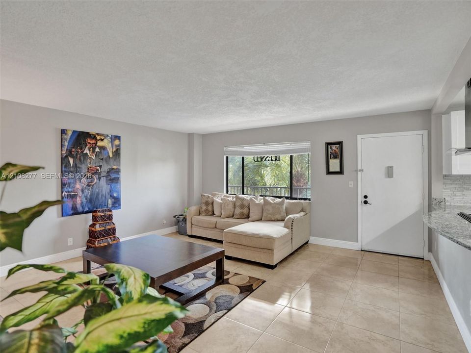 Open-concept interior featuring neutral wall tones, large-format tile flooring, a wide window with blinds, and a stone-surface kitchen counter