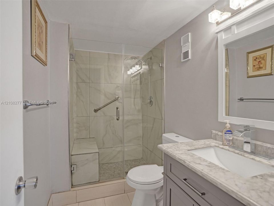 Bathroom featuring a walk-in shower with glass enclosure, built-in tiled bench, stone-finish countertop, vanity with shaker-style cabinetry, and wall-mounted light fixture
