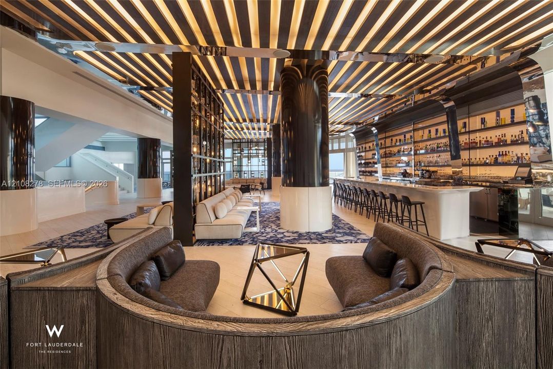 Linear coffered ceiling featuring integrated lighting, custom bar with stone-finish surfacing, curved wood-paneled seating nook, polished architectural columns, and expansive shelving